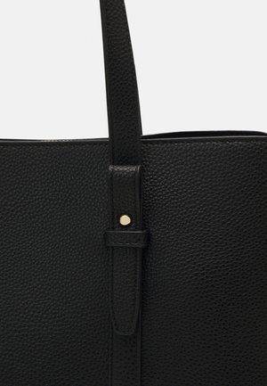 Zbliżenie na pasek torby z czarnej fakturowanej skóry, przymocowany małym, okrągłym, złotym nitem.