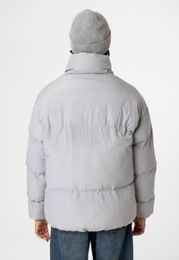 Lichtgrijze pufferjack met een hoge kraag, voorzien van horizontale stiksels en elastische boorden. Draagt over een blauwe spijkerbroek en een grijze gebreide beanie.