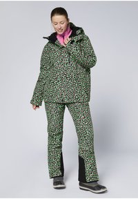 Leopardenmuster-Jacke und Hose in Grün mit pinkfarbenen Akzenten, ausgestattet mit einer schwarzen Kapuze, Reißverschluss und strapazierfähigem Material. Schwarze Schuhe gezeigt.