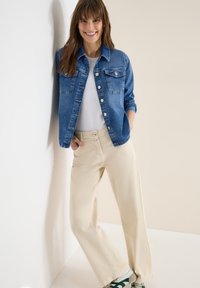 Femme souriante, portant une veste en jean bleue, un haut blanc, un pantalon large beige et des baskets blanches, appuyée contre un mur blanc.