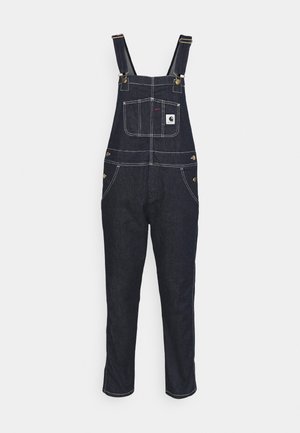 Salopette en denim bleu foncé avec bretelles réglables, poche avant et coutures blanches contrastantes. Comprend du matériel en laiton et des jambes fuselées.