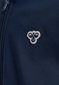 Felpa con zip di colore blu navy caratterizzata da un tessuto texture. Un logo a forma di ape ricamato in bianco e blu scuro è posizionato sulla parte sinistra del petto.