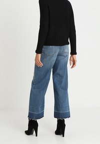 Sininen denim-housut, joissa on korkea vyötärö ja leveät lahkeet, rikotullahelmasta ja erottuvista ompeleista, yhdistettynä tekstuurilla varustettuun mustaan neuleeseen ja mustiin korollisiin saappaisiin.