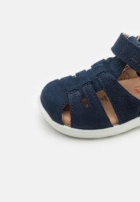 Barnsandaler i marinblått med överdel i mocka, öppen tå-design, kardborreband och vit gummisula. Har utskurna sidodetaljer.