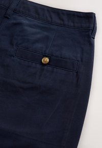 Pantalon bleu marine en tissu lisse, doté d'une poche arrière avec fermeture à bouton doré et détails de coutures visibles.
