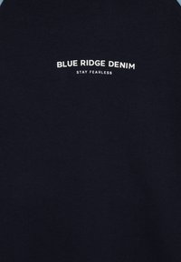 Temno modra tkanina z teksturirano površino, ki vsebuje bel logo z napisom "BLUE RIDGE DENIM" in "STAY FEARLESS" v ukrivljenem dizajnu.