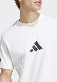 Hvitt bomull t-skjorte med rund hals, med grå aksent og en svart tre-stripes logo på venstre bryst. Korte ermer.