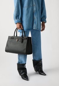 Sort læder håndtaske med en struktureret overflade, dobbelte håndtag, guldfarvet hardware og accentrem. Båret sammen med et blåt denim-outfit.