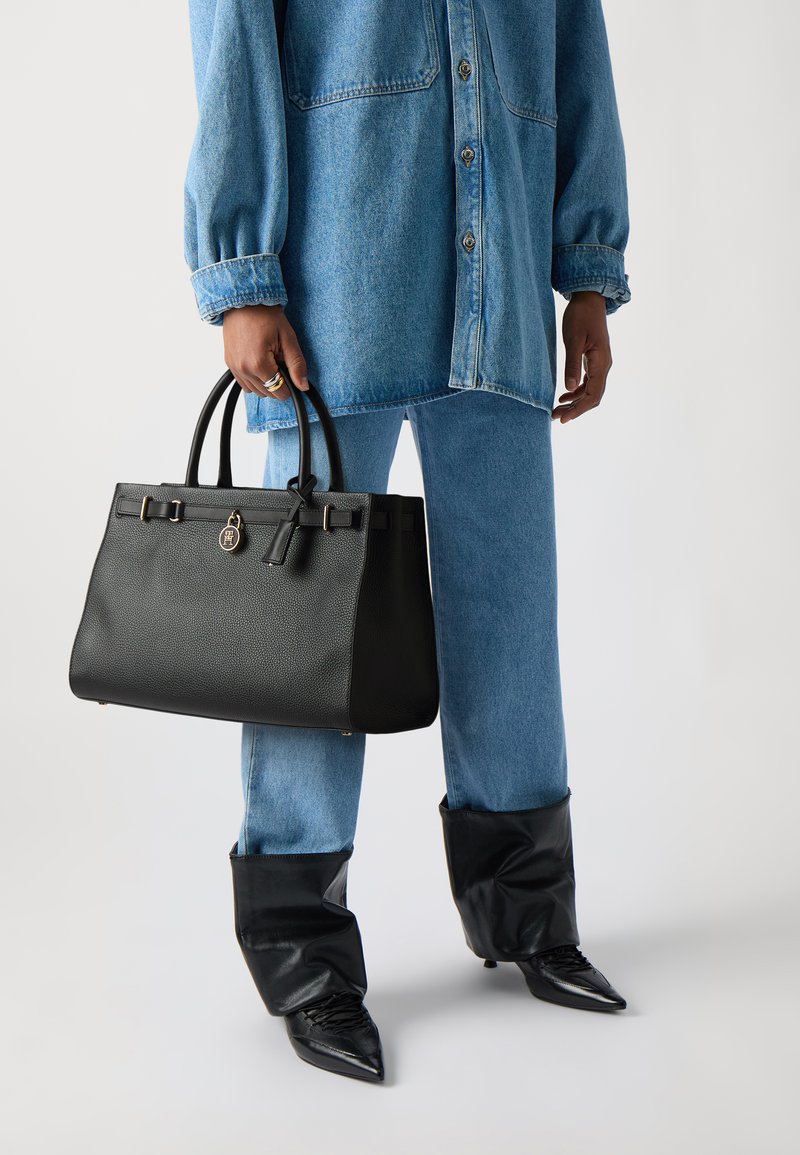 Sac en cuir noir avec une finition texturée, double poignées, quincaillerie dorée et sangle d'accent. Porté avec une tenue en denim bleu.