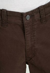 Braune Baumwollhose mit Reißverschluss und Knopfverschluss, ausgestattet mit einem metallischen "Reell"-Akzent und Nähten an Bund und Taschen.
