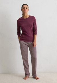 Mujer de pie descalza, vistiendo una blusa de manga larga color burdeos y pantalones rectos de estampado, frente a una pared blanca lisa.