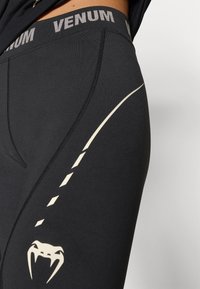 Czarne legginsy sportowe o dopasowanym kroju, wyposażone w kontrastowe jasnobeżowe logo i graficzne akcenty wzdłuż boku i talii.