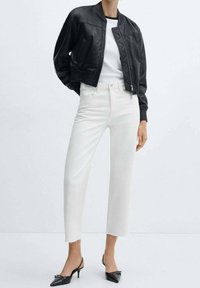 Chaqueta bomber de cuero negro sobre una camiseta blanca de cuello redondo, combinada con jeans blancos cropped y mules negros de punta afilada.