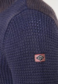 Niebieski sweter z dzianiny z teksturowanej tkaniny, okrągłym wzorem dzianiny oraz małą, czerwoną i pasiastą naszywką z numerem 973 na rękawie.