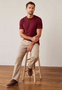 Burgundy crew-neck t-paita, beige suoralahkeiset housut ja ruskeat kengät. Malli istuu puupenkillä, taustalla on sileä vaalea väri.