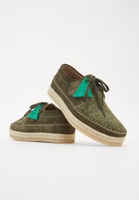 Chaussures en daim vert avec des accents texturés verts, une semelle en jute tressé et un détail de lacet noué. Présente un design unique et des coutures visibles.