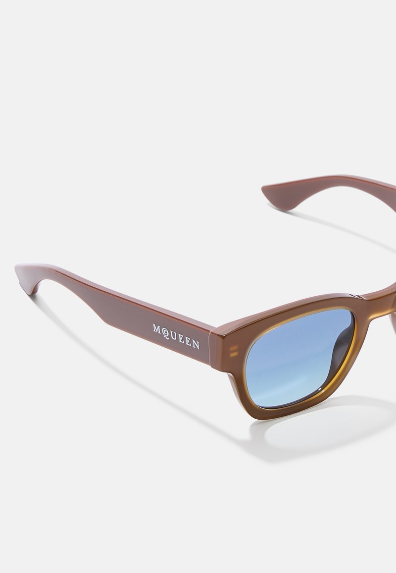 Lunettes de soleil en plastique marron avec des verres teintés en bleu. Elles possèdent une forme de monture carrée et incluent le texte "MQUEEN" sur le côté.