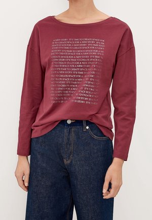 Burgunderfarbenes Langarm-T-Shirt mit rundem Ausschnitt; verfügt über ein wiederholtes Textdesign in Silber. Kombiniert mit dunkelblauen Jeans.