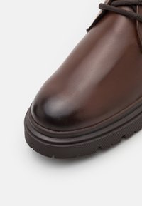Botte en cuir marron avec bout rond, semelle en caoutchouc épaisse et rainurée, et lacets marron foncé sur fond blanc.