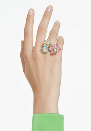 Anillos en forma de flor presentan cristales multicolores en rosa, azul y verde, montados en una banda en tono dorado, llevados en un dedo.