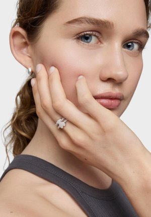 Anillo de plata con un adorno blanco en forma de flor y textura. La mano del modelo descansa sobre su rostro, mostrando una apariencia limpia y natural.