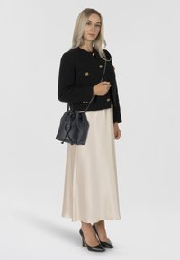 Giacca nera corta con bottoni dorati, vestito di satin color crema, scarpe con tacco nere, mentre tiene una borsa a secchiello blu scuro con chiusura a coulisse.