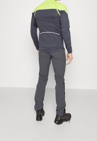 Haut de sport à manches longues en bleu foncé avec un panneau d'épaule vert vif, associé à un pantalon gris, présentant un design ajusté et des accents réfléchissants.