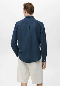 Morsky modrá košeľa s dlhým rukávom, rovný golier, pleat na chrbte a gombíkové manžety. V štýle so svetlými béžovými šortkami.