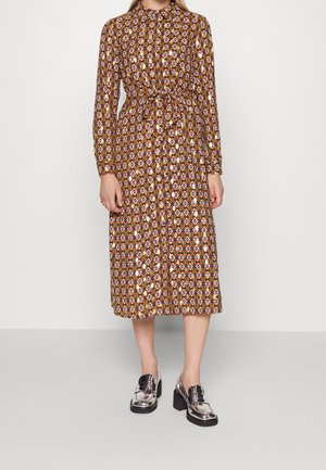 Kobieta ubrana w midi sukienkę z długim rękawem, zapinaną na guziki z przodu, z brązowym geometrycznym wzorem i przewiązanym paskiem w talii, w połączeniu z błyszczącymi srebrnymi butami na szerokim obcasie.