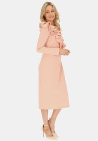 Różowa sukienka midi z długimi rękawami i falbaną przy dekolcie. Gładki materiał z dopasowanym krojem, w połączeniu z cielistymi szpilkami.