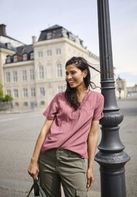 Blouse texturée rose décontractée à manches courtes et décolletée en V, associée à un pantalon cargo vert olive. Fond urbain avec un lampadaire.