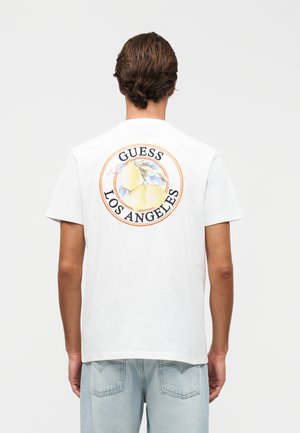 Άνδρας με κοντά καστανά μαλλιά στέκεται με την πλάτη γυρισμένη, φορώντας λευκό T-shirt με γραφικό λεμονιού και το κείμενο «GUESS LOS ANGELES» στην πλάτη.