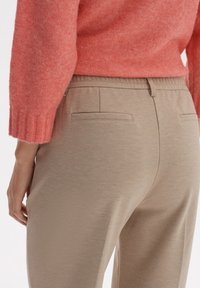 Beige strukturierte Hose mit entspanntem Schnitt, ausgestattet mit zwei Gesäßtaschen und einem elastischen Bund, kombiniert mit einem korallenorangefarbenen Pullover.