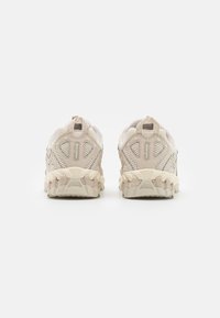 Chaussures de sport beige avec une tige en mesh et des renforcements superposés, une semelle épaisse et texturée, ainsi qu'un col de talon rembourré pour le soutien.