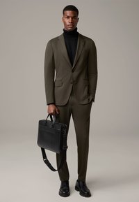 Olivgrüner maßgeschneiderter Anzug mit einem schwarzen Rollkragenpullover, kombiniert mit einer schwarzen Ledertasche und schwarzen Schnürstiefeln. Glatte Stoffstruktur.