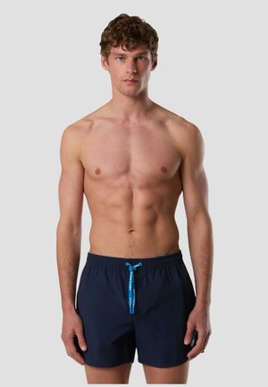 Giovane uomo con capelli ricci che indossa pantaloncini da bagno blu navy con coulisse blu, in piedi davanti a uno sfondo semplice color grigio chiaro.