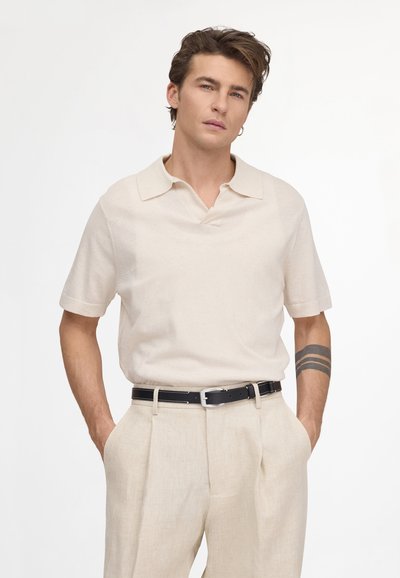Jeune homme aux cheveux châtain clair, portant une chemise beige à manches courtes et un pantalon assorti, avec une ceinture noire et un tatouage sur l'avant-bras gauche.