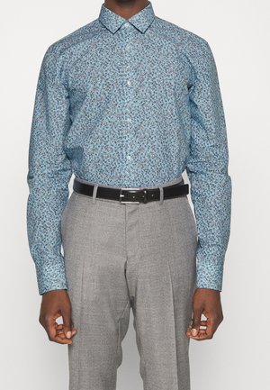 Man wearing light blue floral long-sleeve button-up shirt tucked into gray pleated trousers with a black belt, standing against a white background.