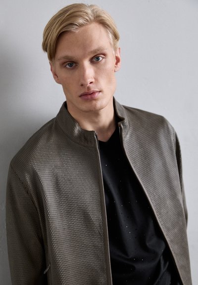 Grå mesh-jakke med oppreist krage, glatt tekstur, glidelås foran, over en svart t-skjorte med subtile prikkete aksenter.