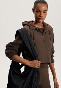 Hoodie corto marrone con cappuccio foderato in pile, abbinato a una borsa nera scura. Il modello tiene la borsa su una spalla, mettendo in risalto le trame sovrapposte.