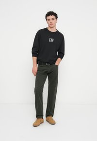 Μαύρο φούτερ με το λογότυπο "GAP", πράσινο παντελόνι κοτλέ και καφέ αθλητικά παπούτσια. Το σύνολο συνδυάζει άνετη εφαρμογή και υφασμάτινες υφές.