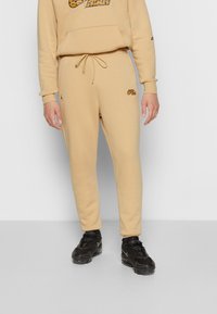 Beiger Hoodie und Jogginghose aus weichem Stoff mit schwarzem und orangefarbenem Grafiklogo. Kombiniert mit schwarzen Sportschuhen.