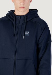 Marineblaue Zip-Hoodie aus weichem Stoff mit Kapuze, Kordeln und Logopatch. Glatte Textur mit kontrastierenden Paneeldetails an den Ärmeln.
