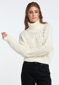 Strickpullover in Off-White mit hohem Rollkragen. Verfügt über strukturierte Muster und gerippte Bündchen, getragen mit schwarzen Jeans.