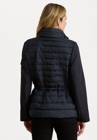 Marine blauwe pufferjack met gequilt textuur, hoge kraag en lange mouwen. Riem aan de taille en details van zijzak met rits zichtbaar.