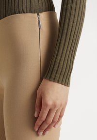 Ribbad olivgrön långärmad topp i kombination med beige stretchbyxor med en synlig sidogenvör och mjuk textur. Handen vilar på byxorna.