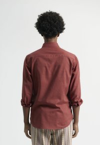 Camisa roja de manga larga hecha de tela tejida, con cuello y mangas arremangadas, vista desde la parte trasera sobre un fondo claro.