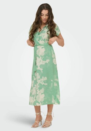 Mujer ajustándose el lazo de la cintura en un vestido midi floral verde y blanco, con sandalias beige de tacón alto y tiras, de pie sobre un fondo blanco liso.