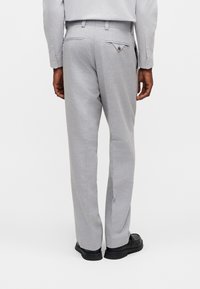 Άνδρας που φορά ανοιχτό γκρι παντελόνι φόρεμα και πουκάμισο, μαύρα παπούτσια, στέκεται με την πλάτη προς το μέρος μας σε απλό φόντο.