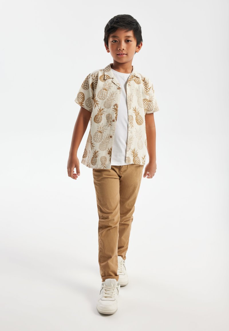 Kortärmad krämfärgad skjorta med brunt ananastryck, i kombination med beige byxor och vita sneakers, på en helt vit bakgrund.